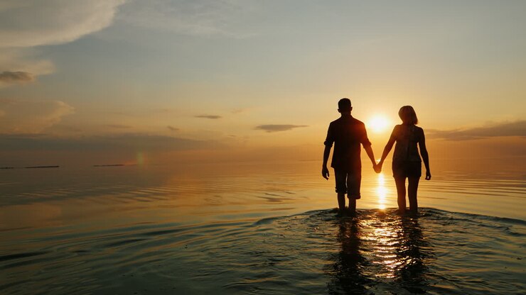 Un homme et une femme vont vers la mer au coucher du soleil en se tenant la main, silhouettes dans la lumière du soleil couchant.