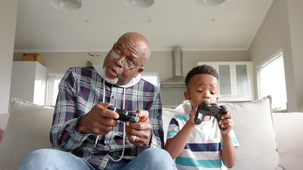 African american grandfather and grandson sitting on sofa and playing video games, slow motion