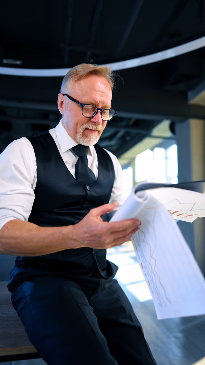 Adult male businessman in white shirt and vest holds the paper with charts. Man shows first graph and thumb up while he reacts negatively on the next graph. Close up. Vertical video.