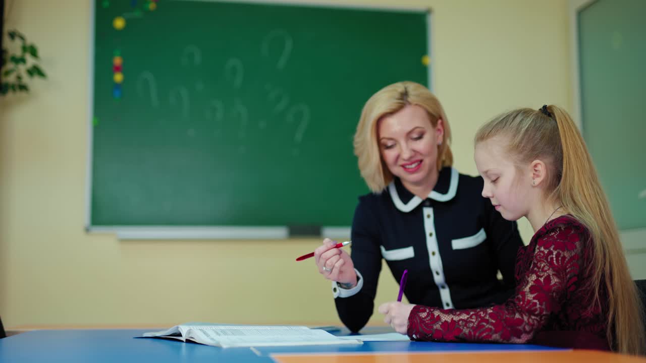 Teacher helps schoolgirl. Little girl and her teacher sitting in the classroom. Woman teaching a clever student on the blackboard background at school.