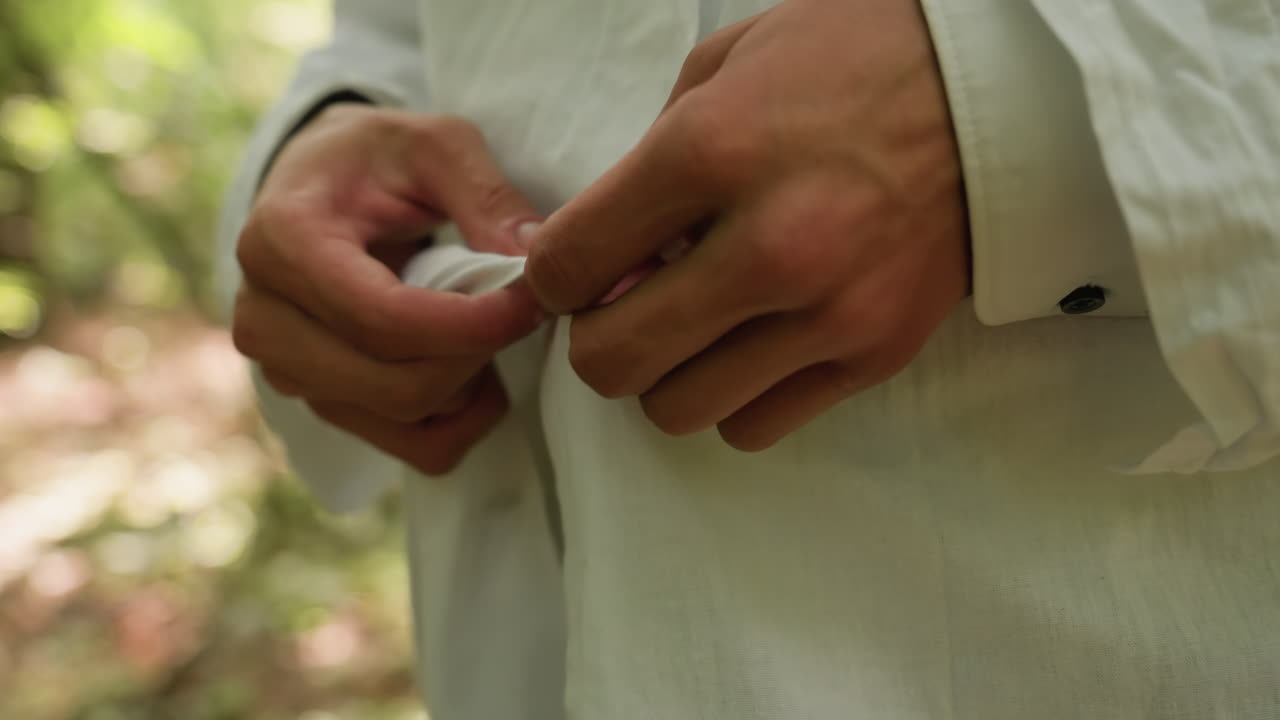 Close up view of young botany student in white shirt opening coat button in blurred forest with bright light, highlighting natural greenery, outdoor preparation, and detailed gesture of concentration