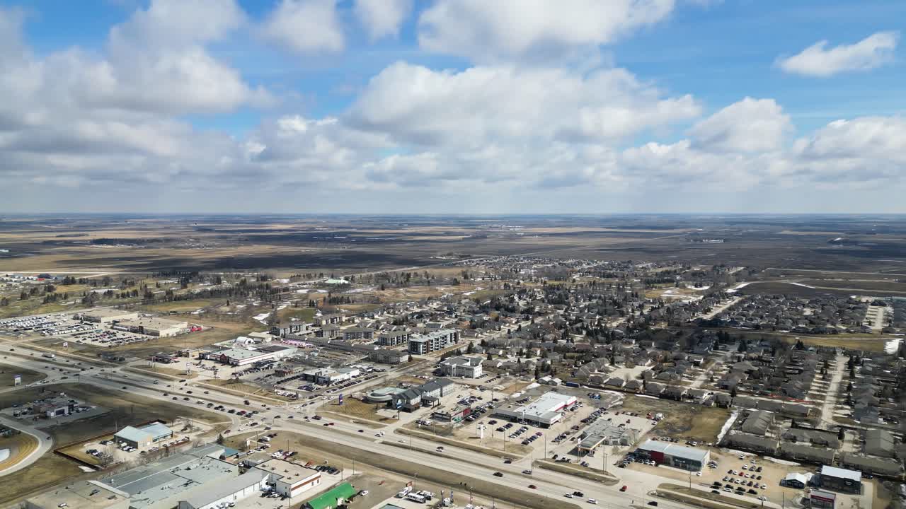 Slow panning aerial drone shot from high altitude showing a small town in southern Manitoba, Canada with distant horizon, small buildings, open space, and surrounding farmland during the daytime