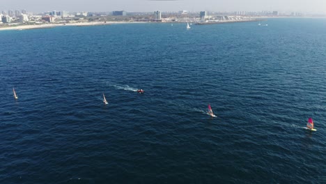 Varios-Windsurfistas-Navegan-A-Gran-Velocidad-Sobre-Las-Hermosas-Aguas-Azules-Del-Mar-Mediterráneo-Con-La-Ciudad-De-Herzeliya-Al-Fondo