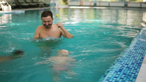 young father is showing to his little son to dive under water in the swimming pool while he is sitting on the border. he is jumping in, swimming, then his mother is lifting him up from the water