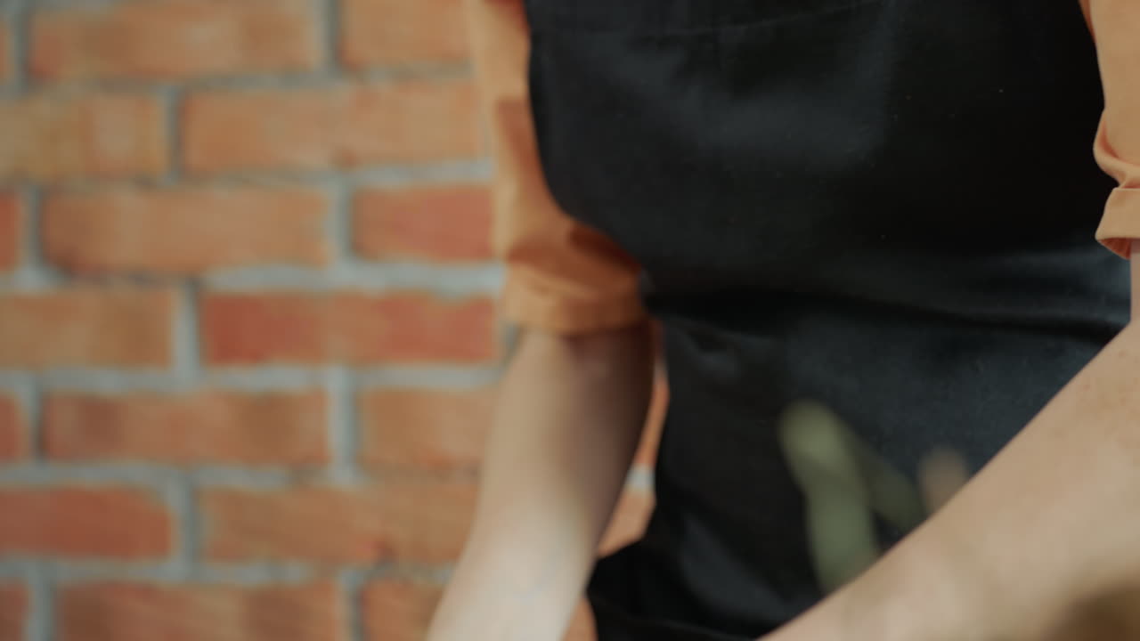 Florist in apron stretches twine while preparing dried grass arrangement on wooden table in rustic studio with brick wall, focusing on handmade natural floral decoration craft using eco friendly materials