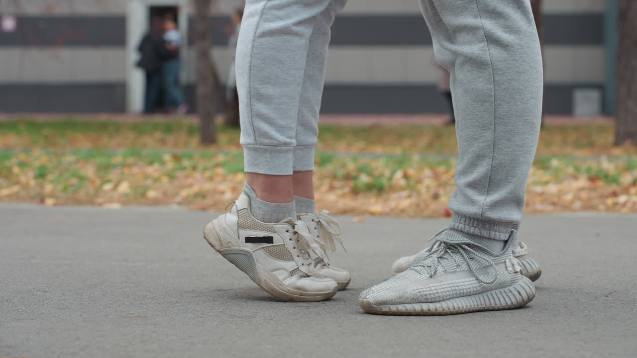 Leg view of two people standing in joggers and sneakers as one person stretches up, lifting feet off ground on paved path with blur background featuring dry autumn foliage and outdoor trees