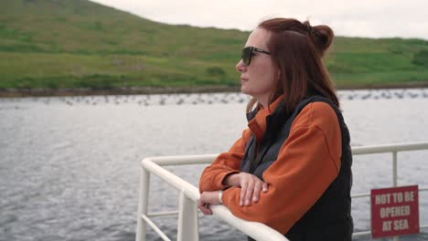 a woman wearing sunglasses enjoys the scenic view from the deck of a ferry or tour boat in ireland. she gazes thoughtfully at the green, rolling hills of the coastline