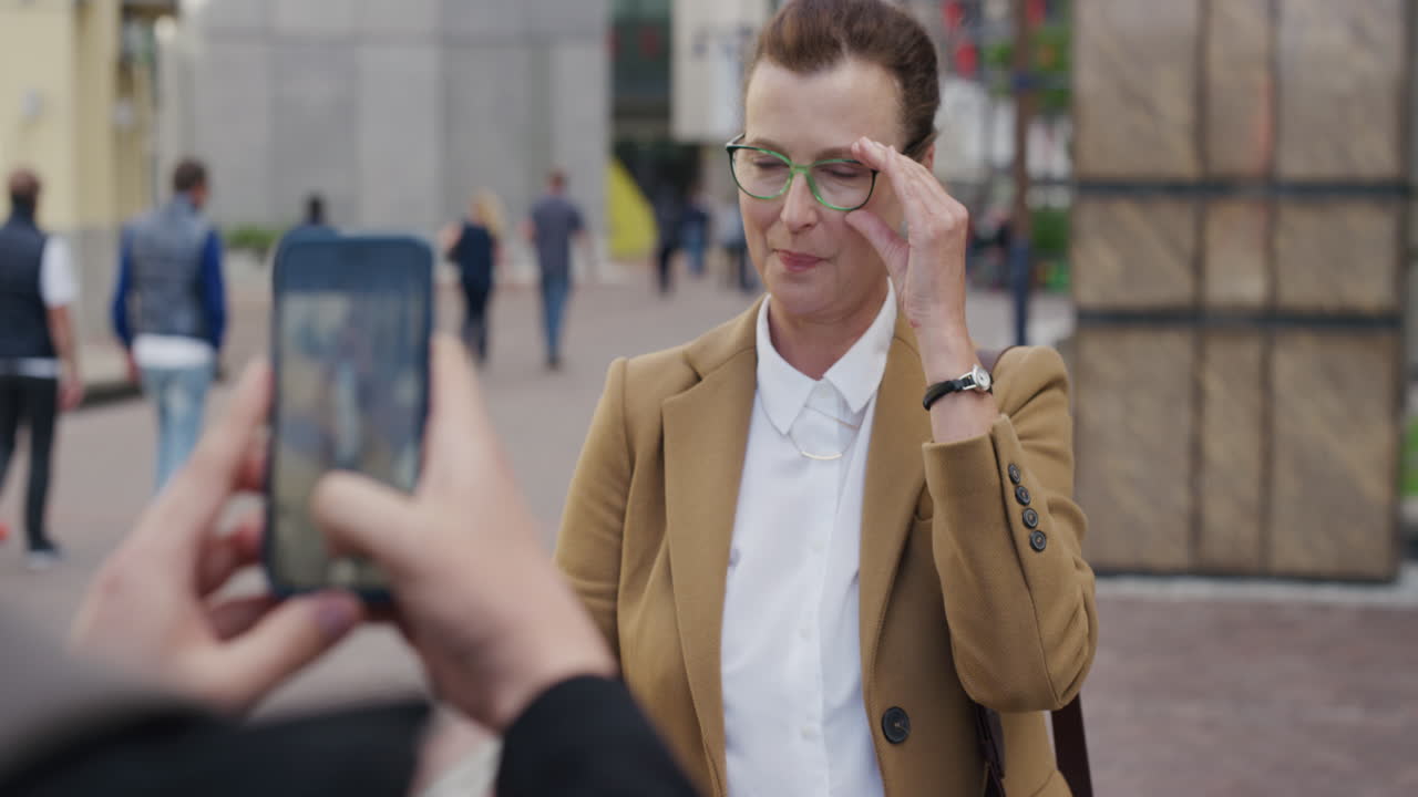 портрет успешной бизнес-женщины среднего возраста, позирующей для фотографии, улыбающейся, наслаждающейся карьерной вехой. мужчина фотографирует с помощью смартфона в городе.