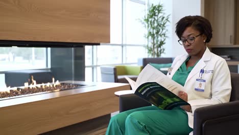 focused health professional reading in a modern lounge near a cozy fireplace, engaged in study with a green medical guidebook in a calm environment