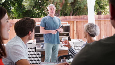 family at a table outdoors turn to dad standing by barbecue