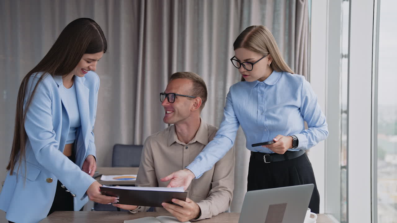 Two female colleagues standing near the man at desk. Teammates speak, look and discuss the documents and laugh a lot.