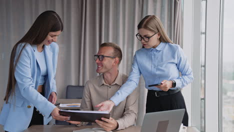 two female colleagues standing near the man at desk. teammates speak, look and discuss the documents and laugh a lot.