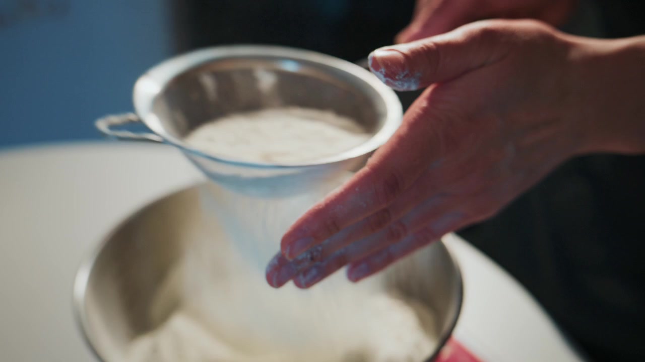 Primo piano di mani che setacciano la farina in una ciotola. Le mani guidano delicatamente la farina verso il basso, mostrando cura. La farina cade dolcemente, creando un effetto di polvere fine. Ideale per la cottura al forno, la cucina. Adatto per ricette.