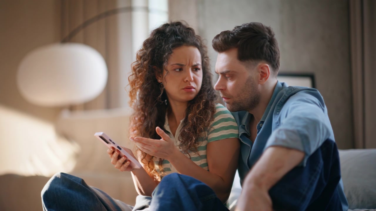 Worried couple looking phone together home. Concerned pair reading bad news.