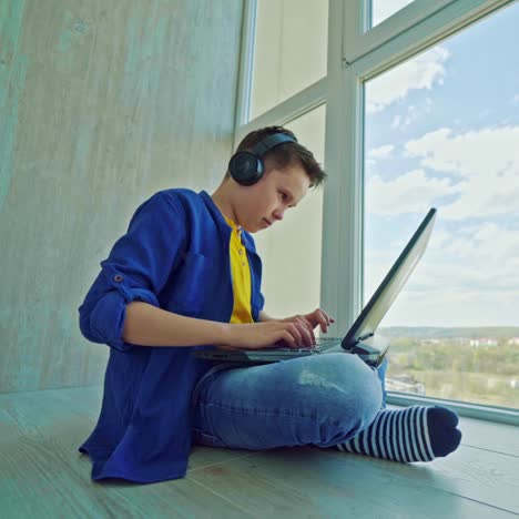happy boy in headphones with a laptop at home. school boy is sitting by the window and playing video games joyfully. gaming concept.