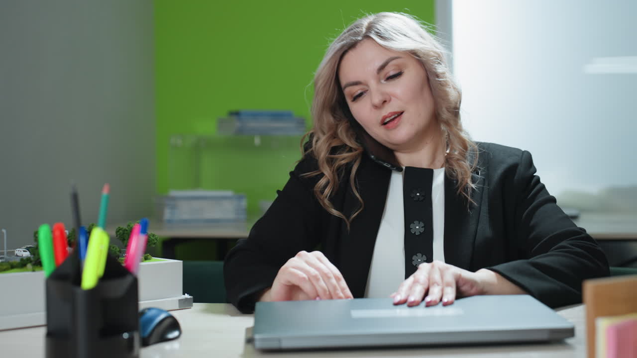 Realtor engaged in phone call sits in office multitasking while opening laptop beside architectural supplies and building model, focused on conversation and project coordination in bright workspace