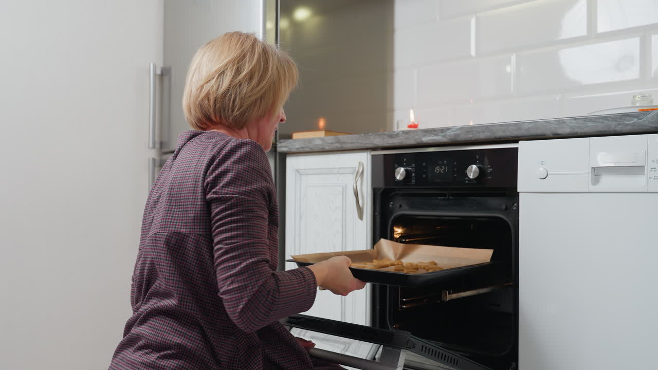 Una mujer saca unas galletas recién horneadas del horno en una cocina moderna, usando una bandeja de horno con galletas. Se ven electrodomésticos, y se prepara para servir o enfriar.