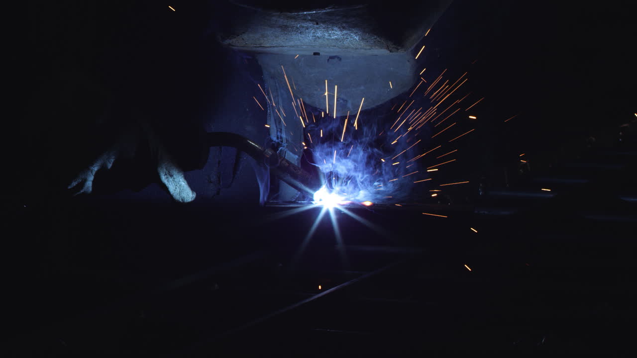 A blacksmith with a welding mask and welders leathers, is welding metal products in the garage, fire sparks fly to the sides. Dark mood. Slow Motion shot with Red Monstro 8K.