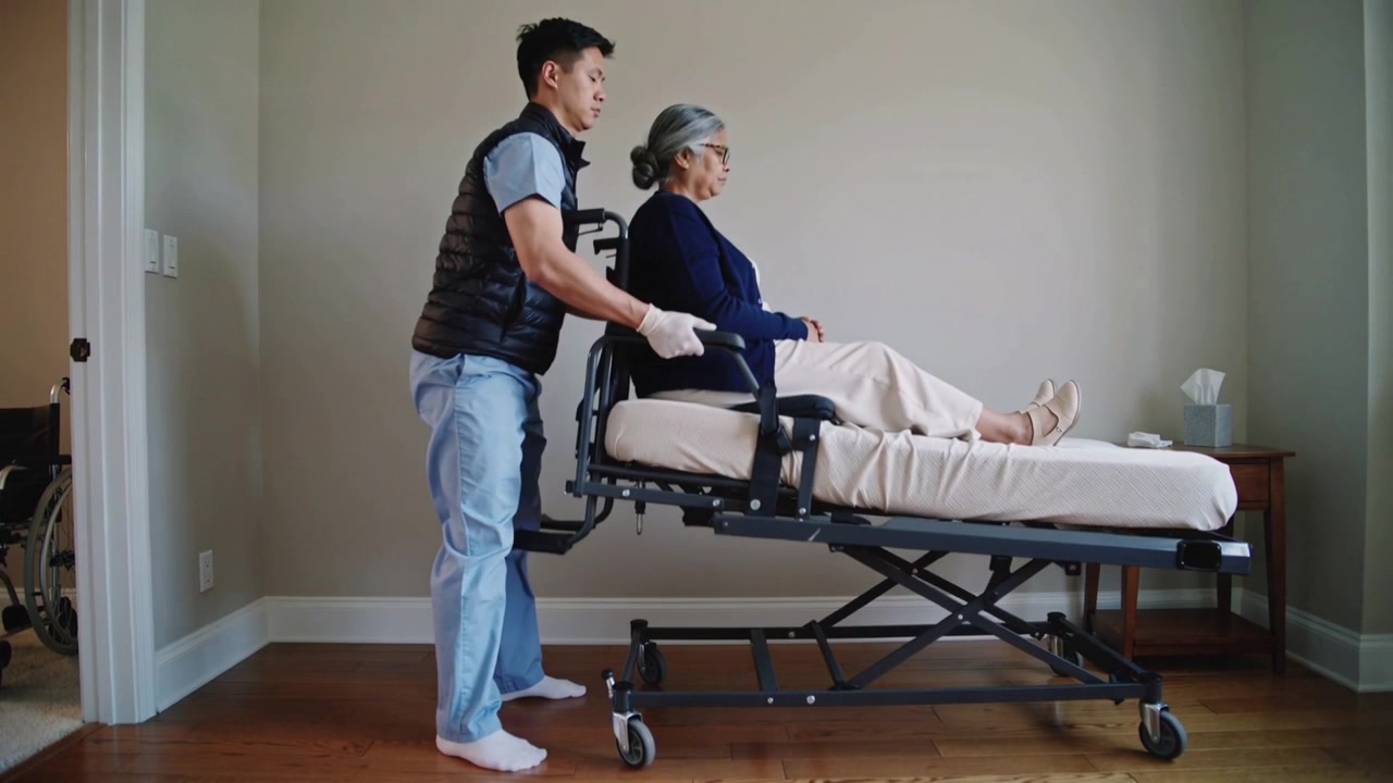 Cuidador con chaleco que se acerca, se pone en cuclillas y levanta a una persona mayor en una cama móvil en casa para realizar una transferencia