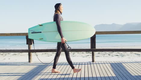 video eines kaukasischen mannes mit dreadlocks in wetsuit, der ein surfbrett trägt und auf der sonnigen promenade spazieren geht