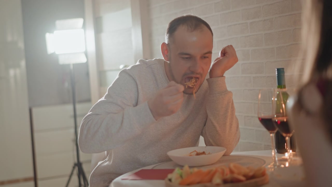 Uomo seduto mangia da una ciotola con un'espressione neutra e concentrata, mentre un compagno sfocato siede di fronte al tavolo in un ambiente da pranzo interno caldo, con morbida retroilluminazione dalla finestra, messa a fuoco superficiale e un'atmosfera sociale calma.