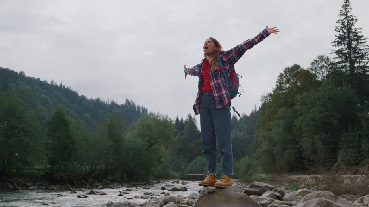 chica feliz de pie en la roca en la orilla del río. excursionista femenina levantando las manos en el aire