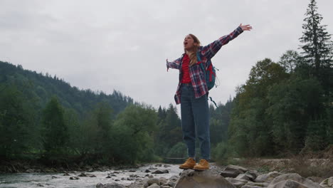 Happy-girl-standing-on-rock-at-river-bank.-Female-hiker-raising-hands-in-air