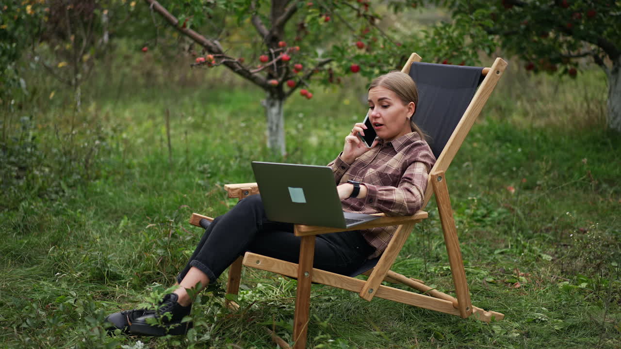 Freelance female specialist sitting in garden chair with laptop and phone. Lady is having conversation over the phone looking at computer screen.