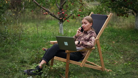 freelance female specialist sitting in garden chair with laptop and phone. lady is having conversation over the phone looking at computer screen.