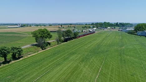 Una-Vista-De-Drones-De-Una-Locomotora-De-Vapor-Con-Vagones-De-Pasajeros-Acercándose-Al-Campo-En-Un-Hermoso-Día-De-Verano