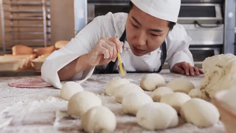 focused asian female baker working in bakery kitchen, cutting dough for rolls in slow motion