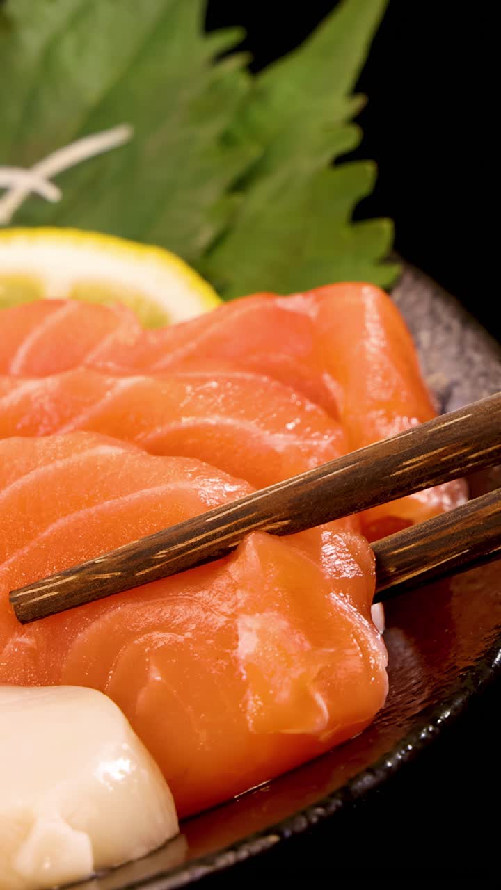 Essstäbchen heben vorsichtig eine Scheibe frisches Lachssashimi von einem traditionellen Sushi-Teller, bei heller Beleuchtung, mit Garnierungen und dunklem Hintergrund