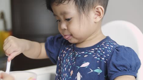 Young-Muslim-Asian-baby-girl-eating-food-using-spoon-at-home-on-baby-chair