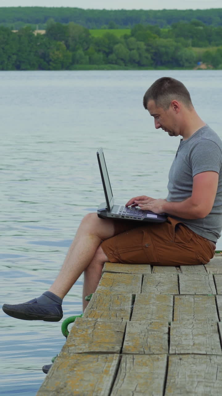 Young man working on laptop while sitting ?n old pier. Vertical video