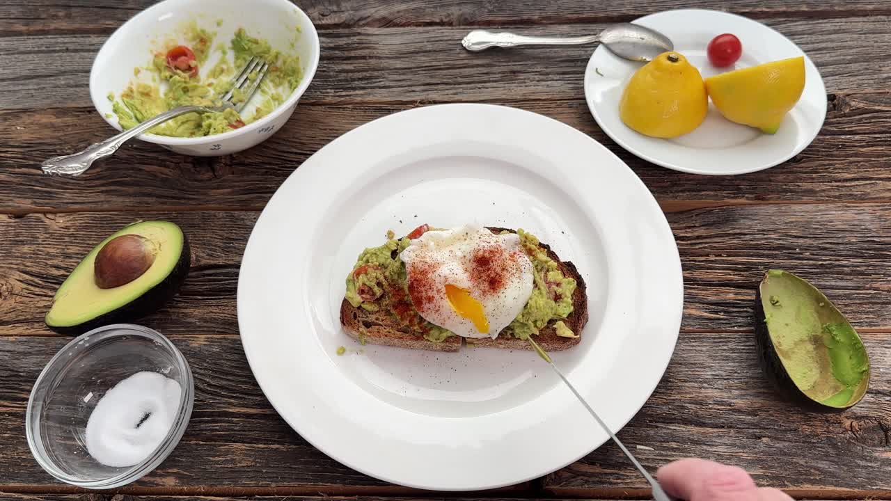 Vista dall'alto di una persona che taglia un uovo in camicia su un avocado toast. Il tuorlo giallo brillante cola sul toast su un tavolo di legno rustico.