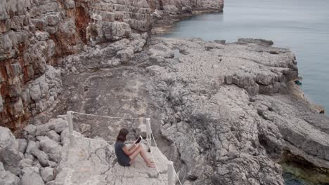 Girl-sitting-on-a-beautiful-rocky-beach-with-tablet-computer-in-her-hand,-wide-shot