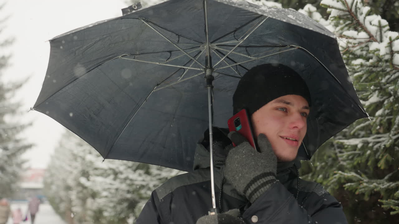 Young white man under umbrella smiling while on phone in snowy park with evergreen trees, wearing beanie and gloves, red handset visible, gentle snowfall, candid student call and winter commute mood