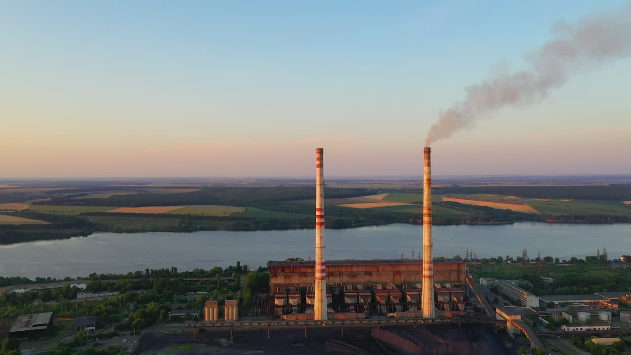 Manufacture near the river. Pipes with dirty smoke on nature background in the evening. Industry among nature at sunset. Aerial view.