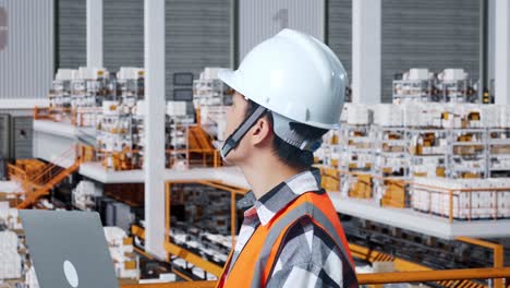 vista lateral de cerca de un ingeniero masculino asiático con casco de seguridad mirando una computadora portátil y mirando a su alrededor mientras está de pie en el almacén con estantes llenos de productos de entrega