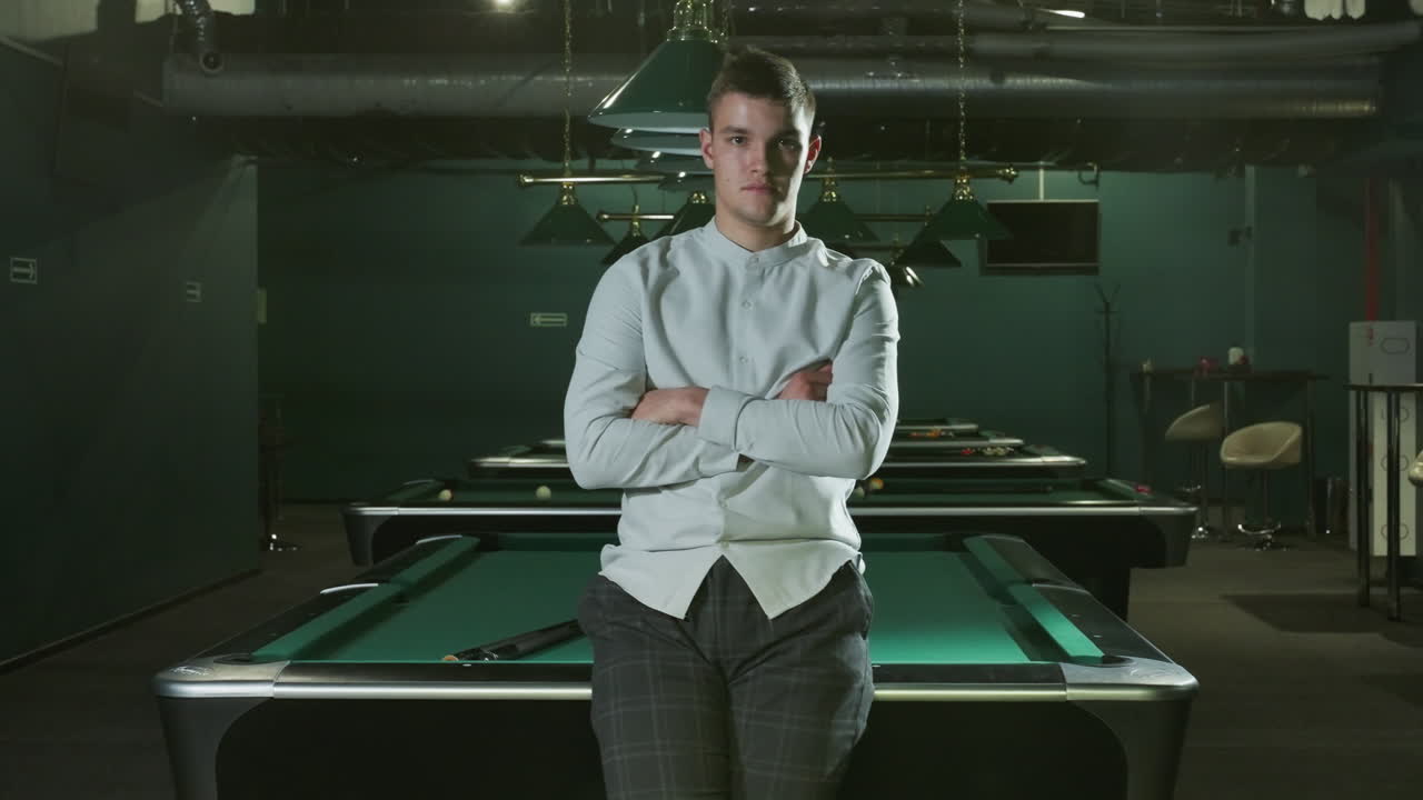 셔츠와 회색 바지를 입은 젊은이가 진지한 얼굴과 팔을 접은 녹색 빌리어드 테이블에 앉아 있습니다. 여러 개의 빌리어트 테이블, 오버헤드 램프, 그리고 현대적인 인테리어가 배경입니다.
