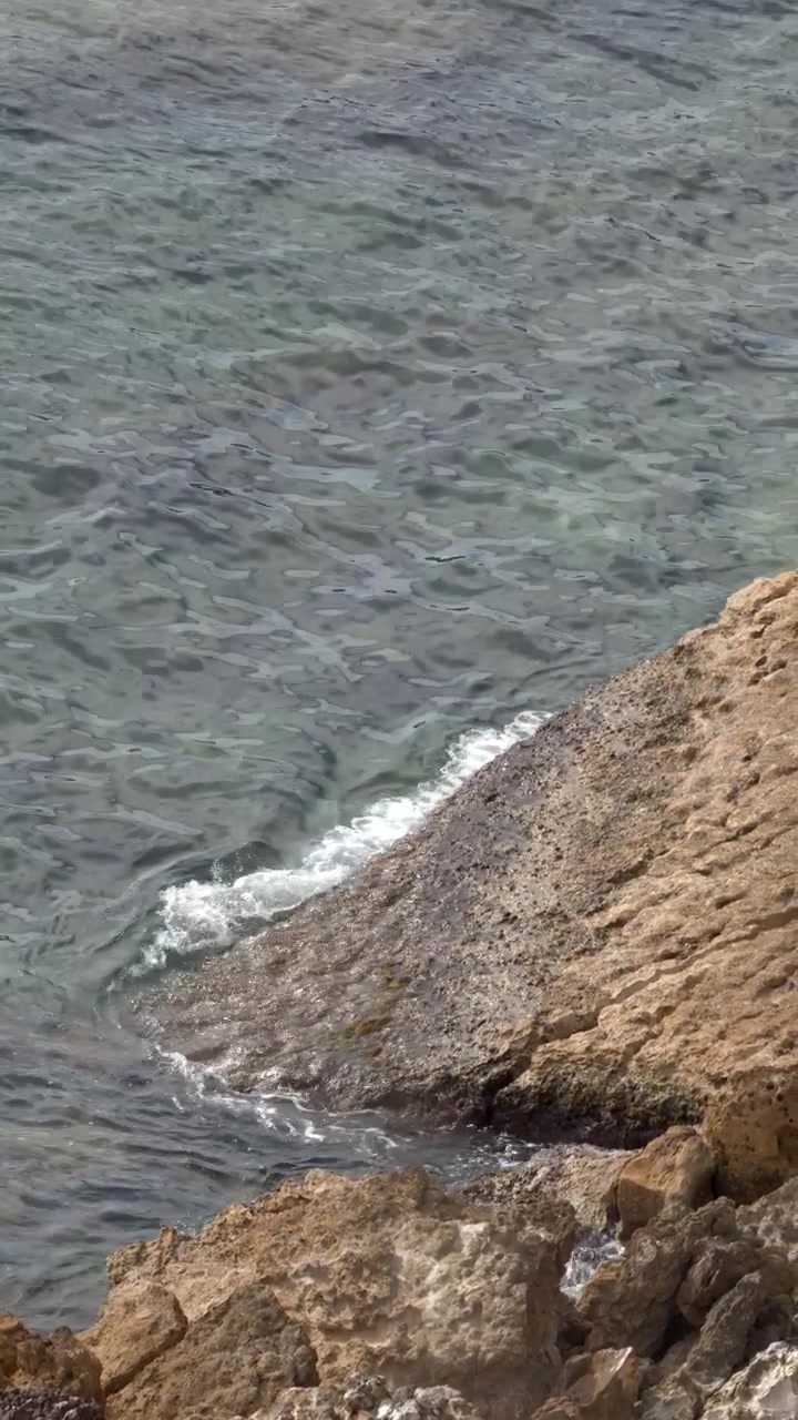 Inquadratura fissa dall'alto di delicate onde mediterranee che si infrangono su una costa rocciosa calcarea frastagliata