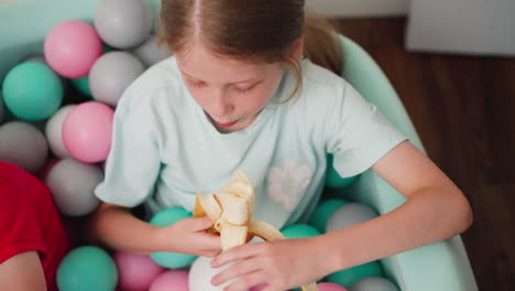 overhead view of little girl eating banana while sitting in colorful ball pit, holding peeled fruit in one hand with partial view of another child in red shirt nearby