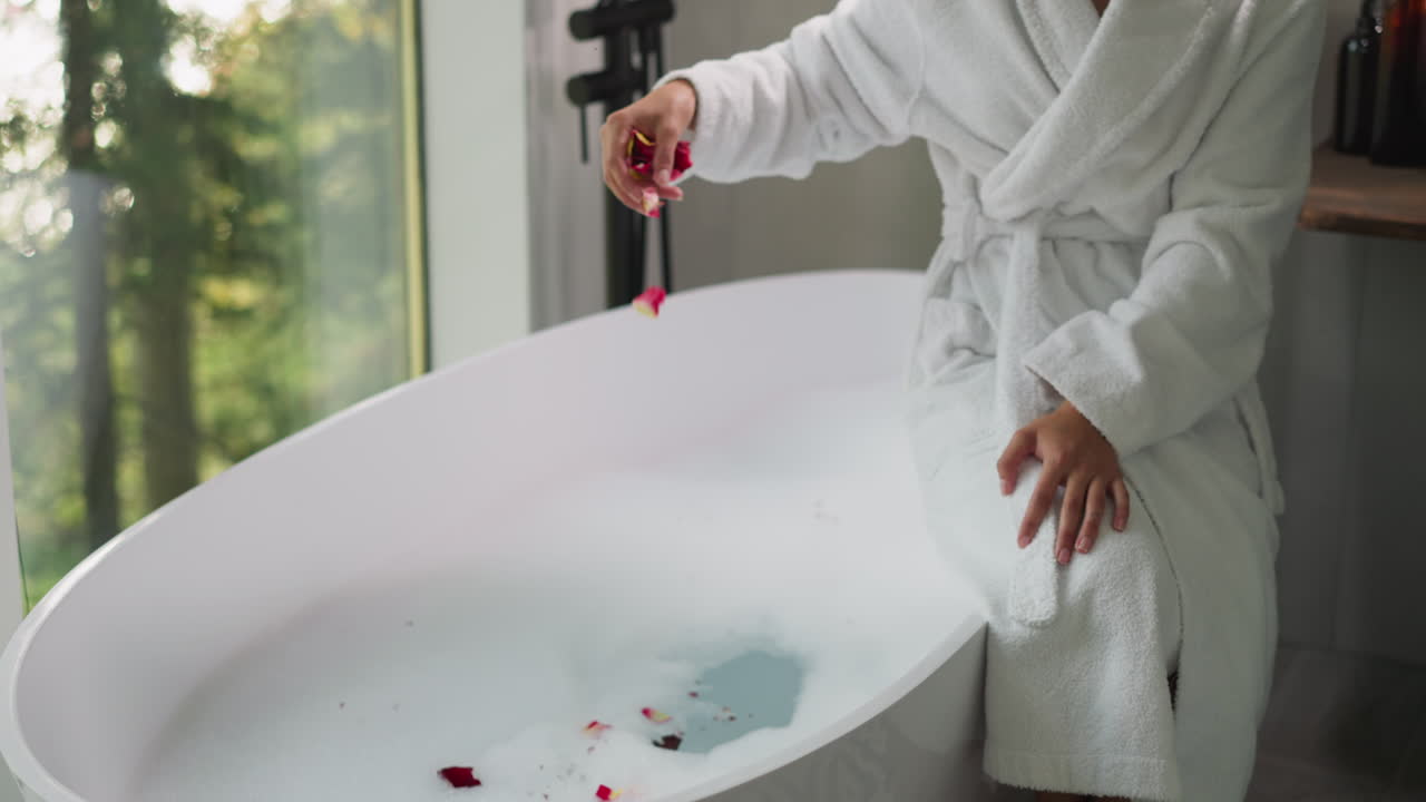 한 여자가 꽃잎을 목욕탕에 던지고, 은 목욕복을 입은 여자가 호텔 방에서 목욕하기 위해 뜨거운 은 물에 꽃을 부어줍니다.