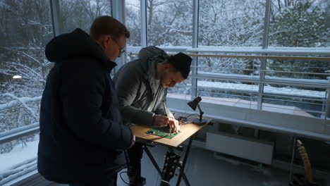 two men collaborating on an electronic circuit board in a modern room with a snowy winter view
