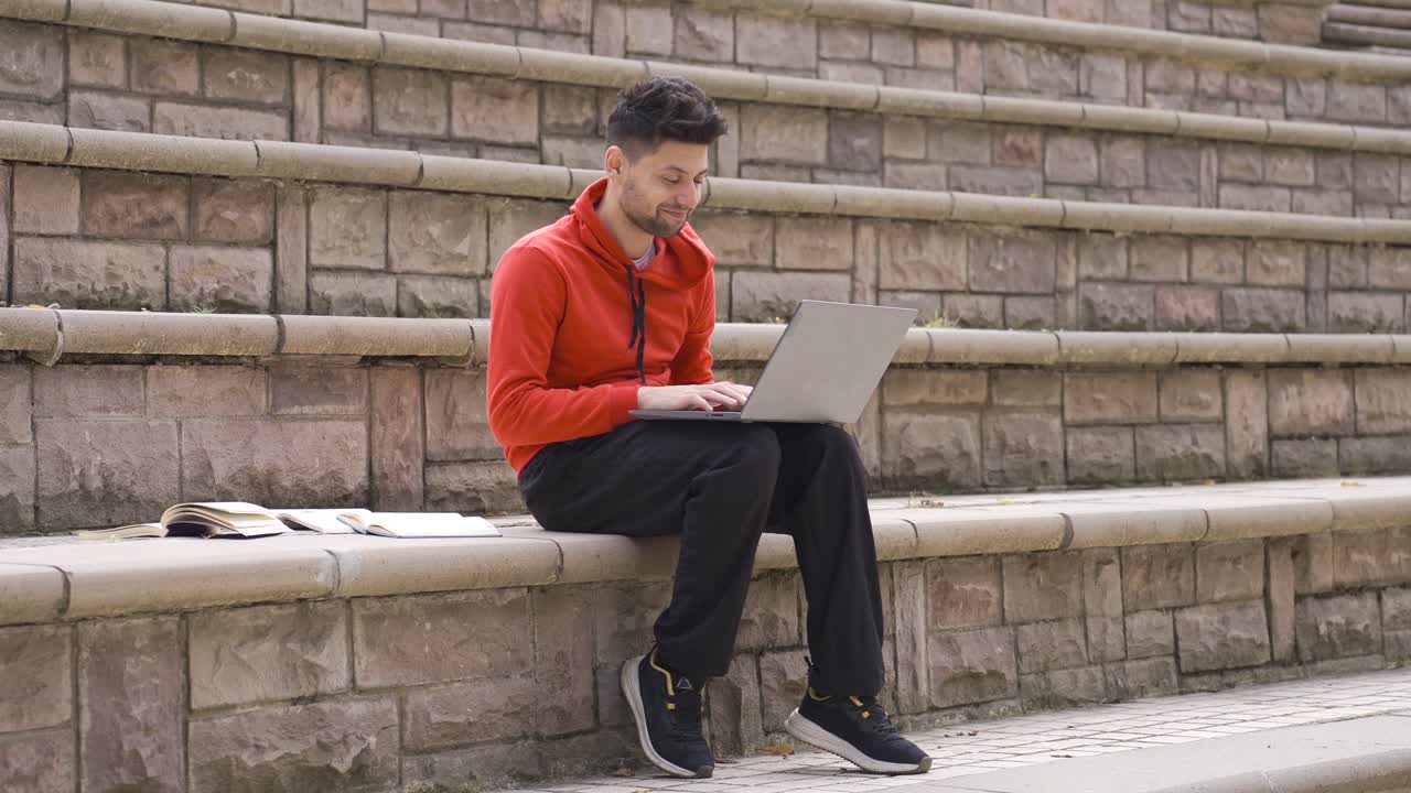 Male student studying outdoors looks at laptop and books.