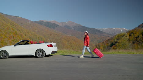 mujer con maleta roja cerca de un coche descapotable blanco en las montañas de otoño