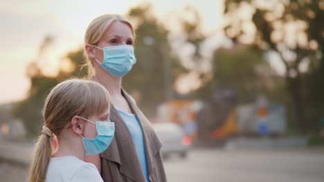 a woman with a child in protective masks are standing near a dirty dusty road in the city