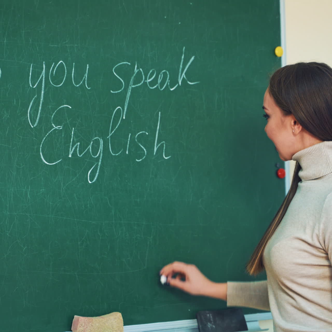 Attractive teacher of English writes on a green board with a chalk. Beautiful long-haired woman is writing question tag on the blackboard and smiles to camera in class.