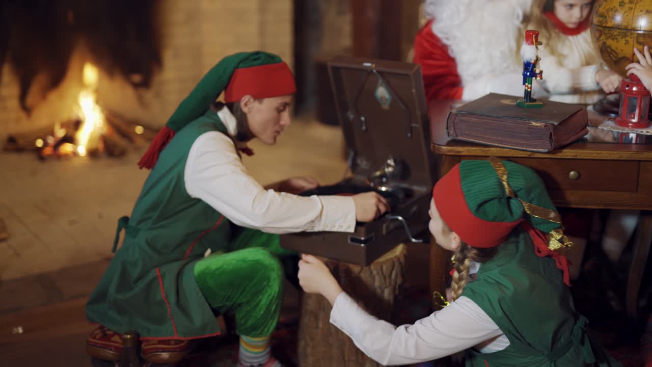 Two elves with old disk in Santa's residence. Happy elves in green costumes putting big disk into old tape recorder.