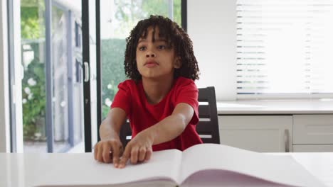 Happy-biracial-boy-sitting-at-table-in-kitchen-reading-braille